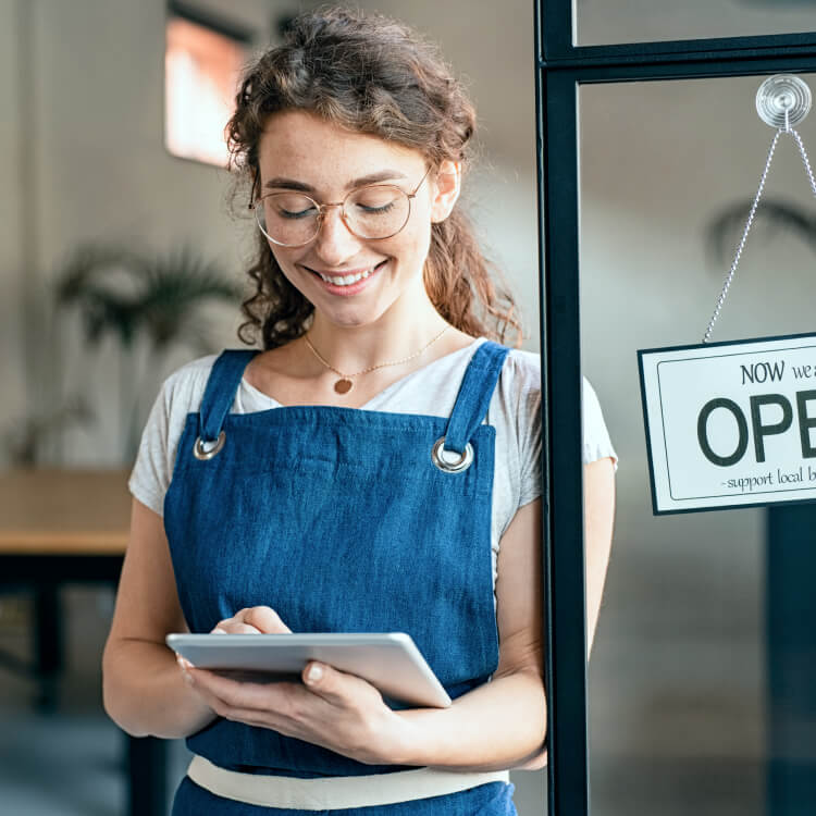 A waitress standing next to a door with a sign that says 'Open'. The waitress is looking down at a tablet and smiling.